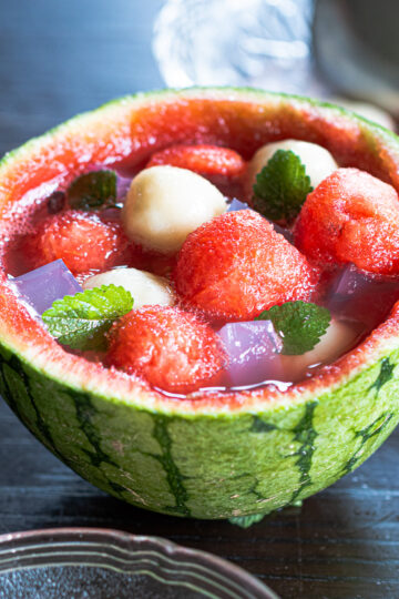 Vegan summer dessert: watermelon fruit punch served in a carved watermelon, topped with chewy banana mochi, pink agar jelly, and fresh mint.