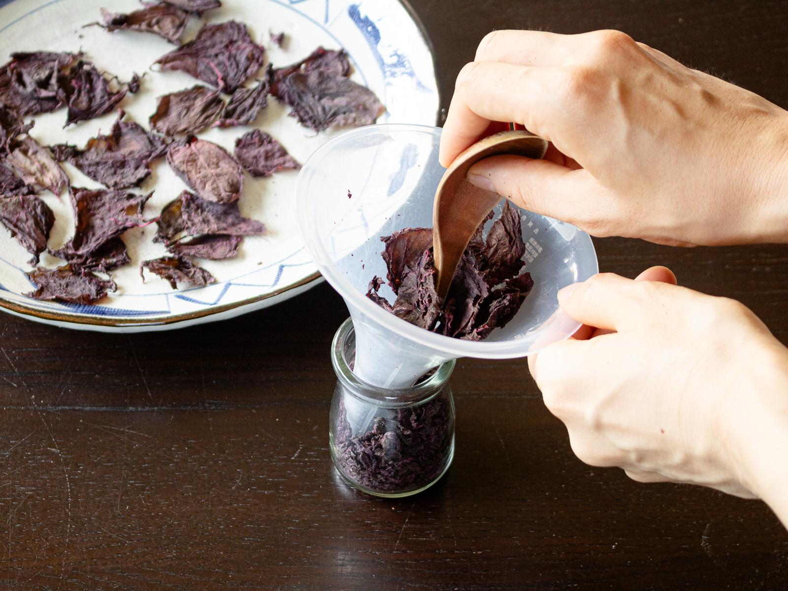 Transferring fully dried red shiso into a jar using a funnel, crushing slightly with a spoon handle or chopstick for fine yukari furikake.