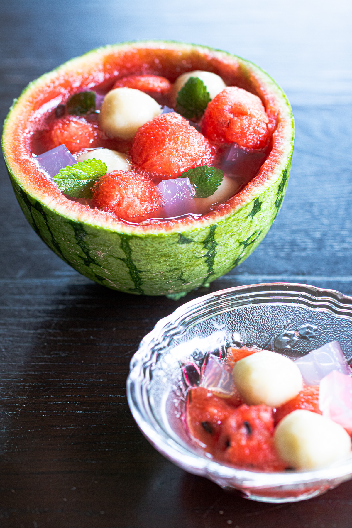 Vegan summer dessert served in a hollowed watermelon bowl, topped with chewy banana mochi dumplings and pale pink agar jelly