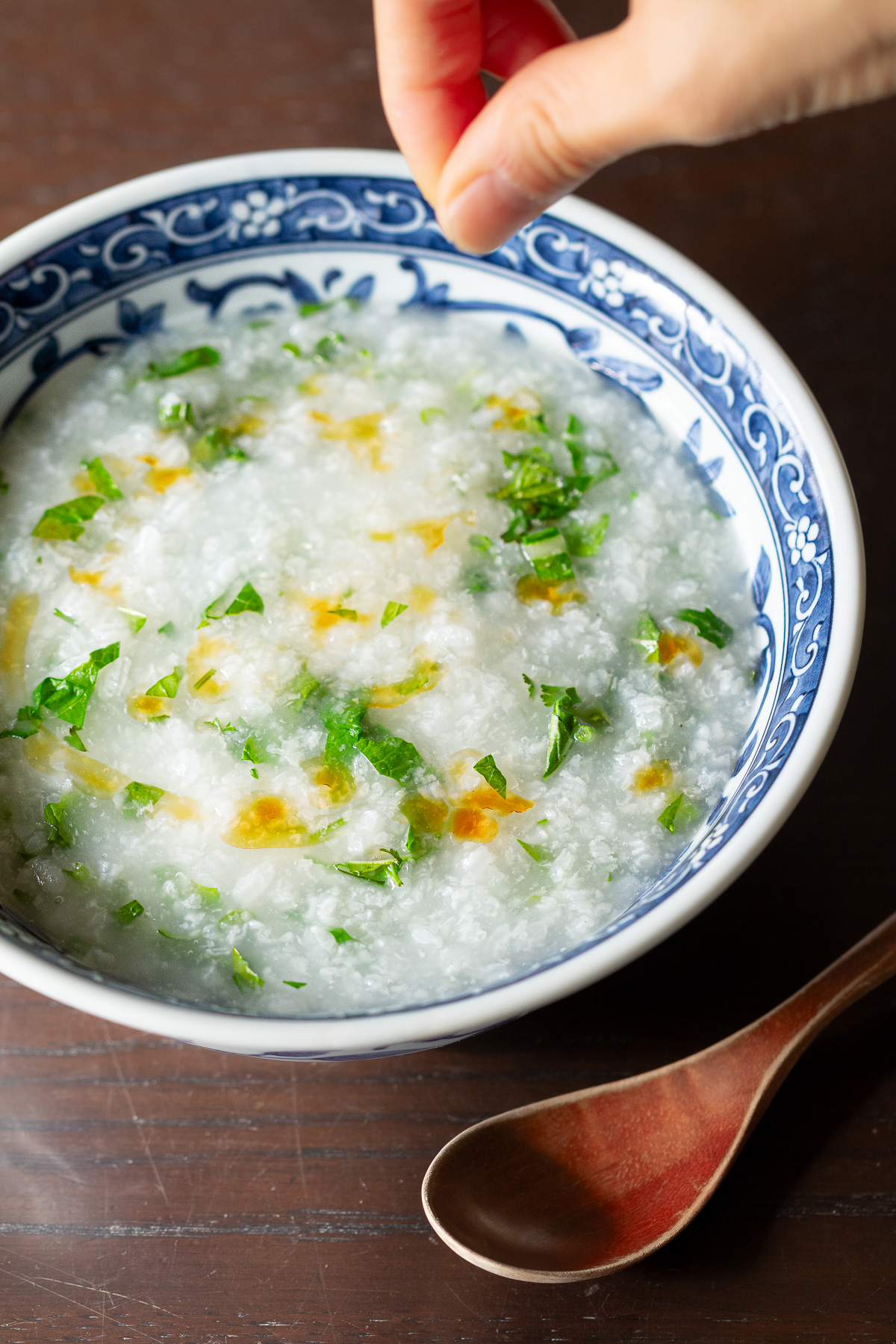 完成したヴィーガン朝粥のクローズアップ - シャキッとした緑の葉野菜、香菜、赤いラー油が青い皿に映える。パラパラと塩をまぶして